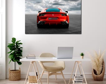 May include: A framed canvas print of a red sports car, viewed from the rear, against a cloudy sky. The car has black exhaust pipes and a small logo. The print is displayed above a white desk with a laptop and a small plant.