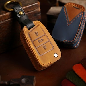 Pode incluir: Uma capa de chaveiro de couro castanho com uma alça de couro preta e um anel de latão. A capa tem símbolos de chave em relevo e um botão branco. Uma capa de couro azul e castanho está no fundo.