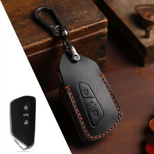 May include: Black leather car key fob cover with orange stitching and a metal carabiner clip. The cover has embossed key symbols and is designed to protect the car key. The key fob is shown next to the key cover.