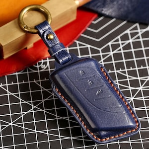 Op de afbeelding: Een marineblauwe lederen autosleutelhouder met een getextureerd oppervlak, contrasterende oranje stiksels langs de randen en een messingkleurige metalen ring bevestigd aan een bijpassende leren riem met een kleine koperen klinknagel.