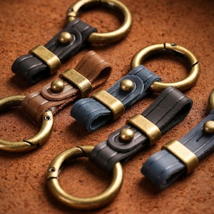 May include: Close-up of several leather keychains in various colors: black, brown, and blue. Each keychain features a brass-colored metal ring and accents. The leather straps are secured with brass-colored rivets and metal clasps.