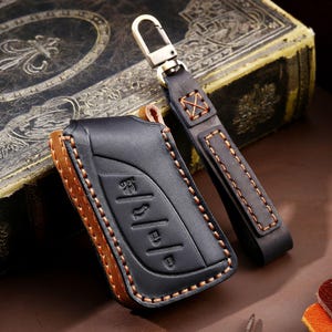 May include: Black leather key fob case with brown trim and stitching. Includes a matching black leather strap with a metal clasp. The case has embossed key symbols. A stylish accessory for car keys.