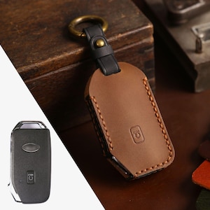 May include: A brown leather key fob cover with a black strap and brass-coloured ring. The cover is stitched with tan thread and features a debossed logo. A black car key is visible on the left.