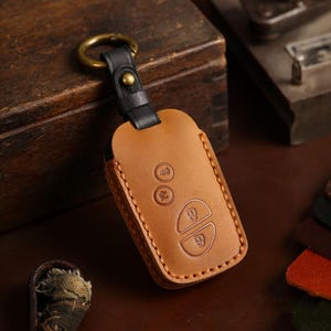 Puede incluir: Una funda para llavero de cuero marrón claro con una correa negra y un anillo dorado. La funda tiene símbolos de llave en relieve y un borde cosido. El fondo es una superficie de madera oscura.