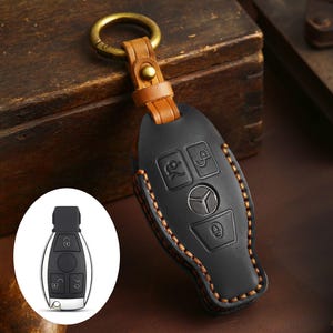 Puede incluir: Funda de llavero de cuero negro con costuras naranjas y un anillo dorado. La funda presenta el logotipo de Mercedes-Benz y los símbolos de las funciones de la llave. Una pequeña llave plateada se muestra en la esquina inferior izquierda.