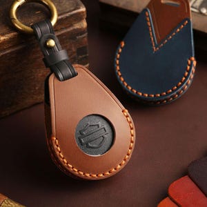 May include: Brown leather key fob with a black Harley-Davidson logo, a black strap, and a gold-colored ring. The fob is stitched with orange thread. Another blue and brown leather key fob is in the background.