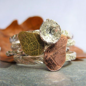 May include: A silver ring with three textured metal pieces. The pieces are in the shape of leaves and a circle. The leaves are gold and copper colored.