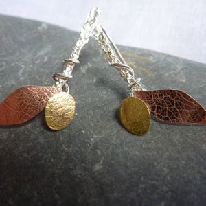 Pode incluir: Um par de brincos de prata com detalhes em folha de cobre e ouro. Os brincos são feitos de fio de prata torcido e apresentam uma folha de cobre texturizada com um círculo de ouro preso a cada um.