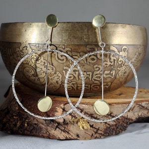 Puede incluir: Un par de pendientes de aro de plata con un acabado texturizado. Cada pendiente tiene un pequeño disco dorado que cuelga de una fina varilla de plata.