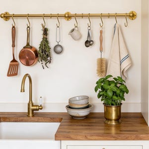 Puede incluir: Escena de cocina con grifo de latón, fregadero blanco y encimera de madera. Una olla de latón con albahaca junto a una pila de cuencos. Colgados, utensilios de cocina, una sartén de cobre y un paño de cocina.