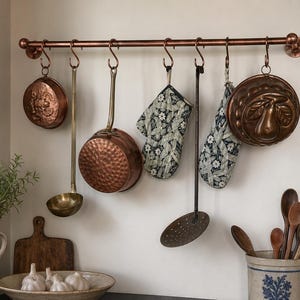 Puede incluir: Una barra de cocina de cobre con utensilios de cocina y guantes de horno colgantes. Los artículos incluyen una sartén redonda en relieve, una olla de cobre martillado, un cucharón, una espumadera y un molde decorativo. Los guantes de horno tienen un estampado floral. Cucharas de madera en un tarro de cerámica.