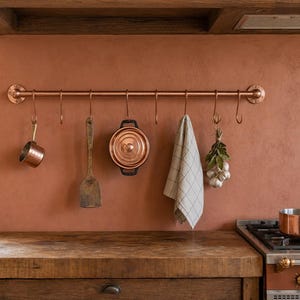 May include: A copper pot rack with hanging kitchen utensils and cookware. The rack holds a small copper saucepan, a wooden spatula, a copper pot, a checkered towel, and a bundle of garlic. The kitchen has a copper-colored wall and a wooden countertop.