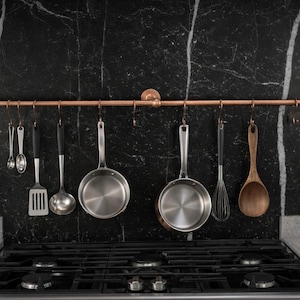 Puede incluir: Un estante de ollas de color cobre con varios utensilios de cocina colgando. Se ven ollas de acero inoxidable, un batidor y cucharas de madera. El estante está montado en un salpicadero de mármol negro sobre una cocina de gas.