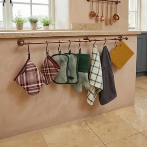 Puede incluir: Una barra de cocina de color cobre exhibe varios textiles de cocina. Los artículos incluyen manoplas a cuadros y a rayas, un paño de cocina a cuadros, una toalla gris y un salvamanteles amarillo. Los artículos cuelgan de ganchos.