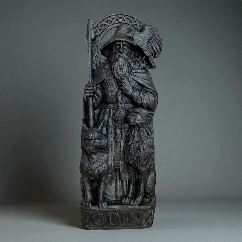 Peut inclure: Une statue gris fonc&eacute; d'Odin, une figure avec une longue barbe, un chapeau et une lance. La statue repr&eacute;sente un corbeau, deux figures animales et le mot "ODIN" &agrave; la base. La statue pr&eacute;sente des d&eacute;tails complexes et un aspect vieilli.