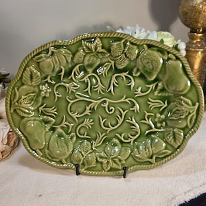 May include: Green ceramic serving platter with embossed fruit and vine design. The oval-shaped platter features pears, strawberries, and other fruits, with a rope-like border. The platter is displayed on a black metal stand.