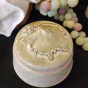 May include: A round, light green ceramic jar with a lid featuring a raised grape leaf and grape cluster design. The jar has a light beige rim and a slightly textured surface. The background includes a white object and a bunch of decorative grapes.