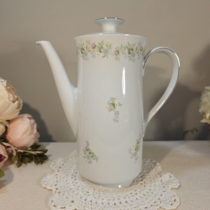 May include: White porcelain coffee pot with a floral design. The pot has a tall, slender shape with a curved spout and handle. Delicate floral patterns in pastel colors adorn the top and body. The lid and handle are trimmed with silver.