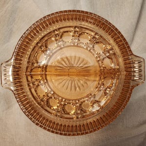 May include: A vintage, amber-colored glass divided serving dish. The round dish features a central sunburst design, geometric patterns, and two handles. The dish is divided into two sections and is ideal for serving snacks or appetizers.