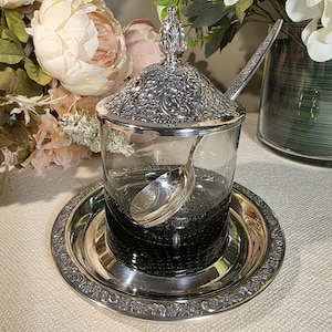 May include: A silver-plated sugar bowl with a glass interior, a matching lid, and a small spoon. The bowl sits on a silver-plated tray. The lid and tray have ornate floral patterns. The bowl is filled with a dark substance.