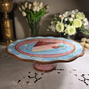 May include: A decorative cake stand with a scalloped edge. The plate features a blue and pink design with a slice of raspberry mousse cake. The base is pink with brown polka dots. The text "Raspberry Mousse" is written on the plate.