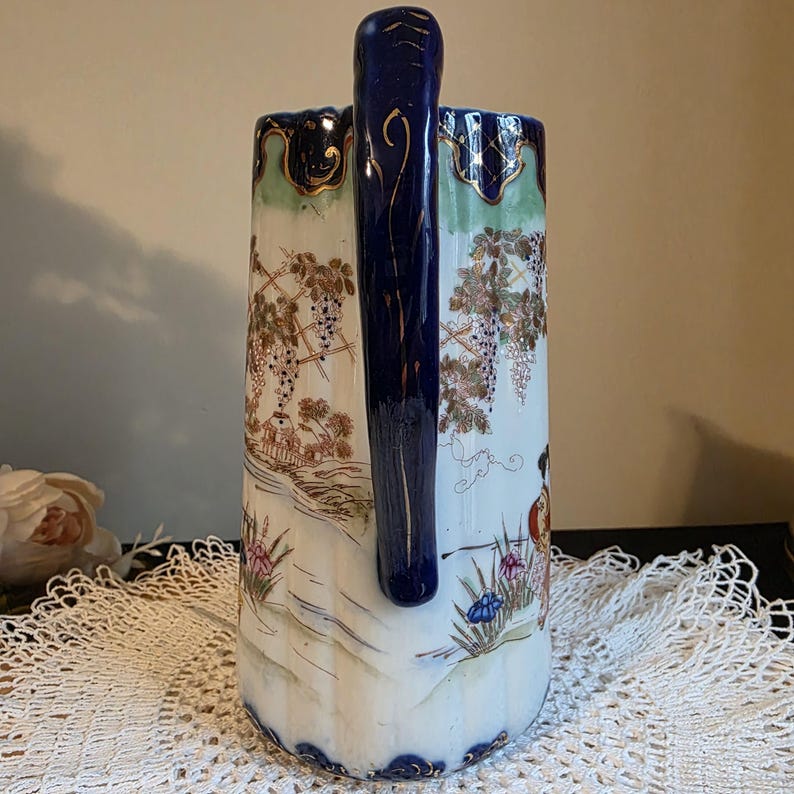 May include: A tall, ornate ceramic pitcher with a dark blue handle and rim. The pitcher features a painted landscape scene with grape vines, figures, and a river. Gold accents adorn the rim and handle, adding a touch of elegance to the design.