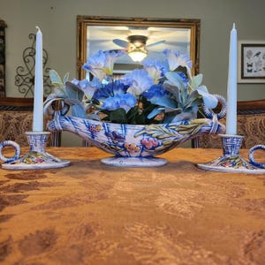 May include: A decorative centerpiece featuring a floral arrangement of blue flowers and greenery in a ceramic bowl. Two matching candle holders with white candles flank the bowl. The bowl and candle holders have a blue and white floral design.
