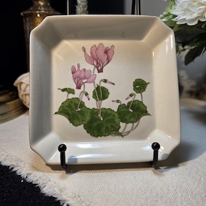 May include: Square, off-white decorative plate with a floral design. The plate features a pink cyclamen flower with green leaves. The plate is supported by a black metal stand, and has a slightly cracked surface.