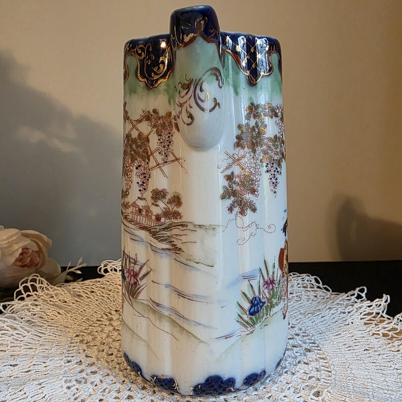 May include: A tall, white ceramic pitcher with a detailed landscape design featuring grape vines, houses, and a river. The pitcher has a blue rim and handle, with gold accents. The design is hand-painted.