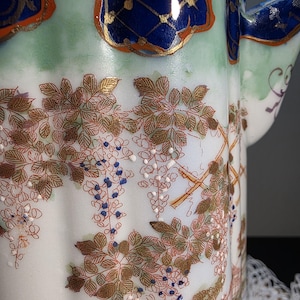May include: Close-up of a white porcelain teapot with a floral design. The teapot features a detailed pattern of brown leaves, blue and white flowers, and gold accents. The top rim is decorated with blue and gold patterns.
