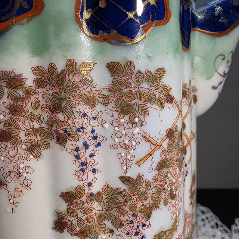 May include: Close-up of a white porcelain teapot with a floral design. The teapot features a detailed pattern of brown leaves, blue and white flowers, and gold accents. The top rim is decorated with blue and gold patterns.