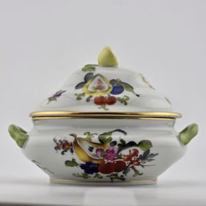 May include: A white porcelain lidded tureen featuring hand-painted fruit motifs including pears, grapes, and berries in vibrant colors. The bowl has two light green handles, a gold-trimmed rim, and a small yellow finial on the domed lid.