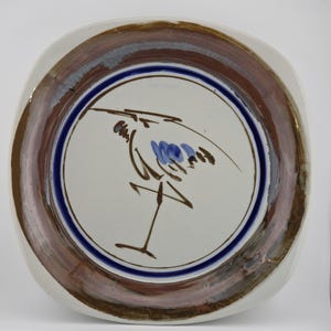 May include: A square ceramic plate with rounded corners featuring a central hand-painted brown line drawing of a bird with blue accents. The plate has a wide, textured border in shades of brown, tan, and grey, framed by a thin, dark blue circular line.