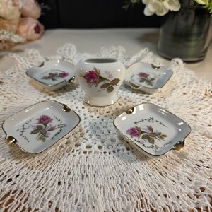 May include: A vintage porcelain set featuring a small vase and four ashtrays. Each piece is white with a gold trim and a floral design of pink roses and green leaves. The ashtrays have a square shape with rounded corners.