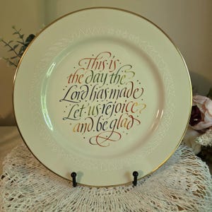 May include: A round cream-colored ceramic plate with a gold rim. The center features decorative calligraphy in red, green, and gold ink that reads, This is the day the Lord has made. Let us rejoice and be glad. The border has white line art.