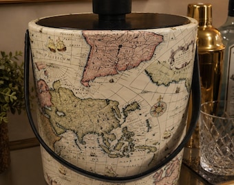 May include: An ice bucket with a vintage map design. The bucket has a black handle and lid, and the map features continents in shades of pink, green, and beige. The bucket is cylindrical and sits on a mirrored surface.