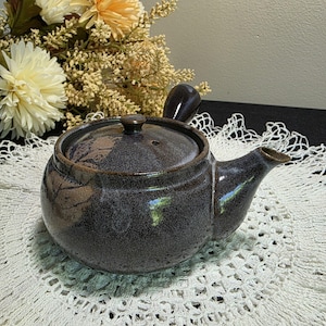 May include: A dark brown ceramic teapot with a rounded body, a side spout, and a small handle on top. The teapot has a lid with a small knob and is placed on a white lace doily. Dried flowers are in the background.