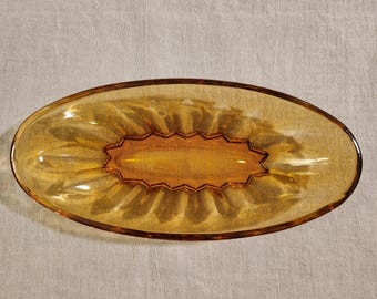 May include: An amber-coloured, oval-shaped glass dish. The dish has a decorative, ridged design around the edges and a detailed, rectangular centre. The glass has a glossy finish, reflecting light. The dish is likely used for serving or display.