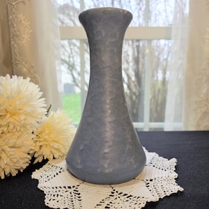 May include: A blue-gray vase with a textured surface. The vase has a flared top and a rounded base. It sits on a white, crocheted doily. Artificial yellow flowers are to the left of the vase.