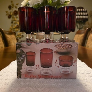 May include: Three ruby red glass goblets with clear stems and bases are positioned atop their original cardboard packaging. The box features printed images of various ruby stemware pieces and the text Ruby Stemware in red lettering.