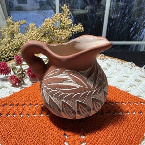 May include: A small, terracotta-colored pitcher with a handle and a spout. The pitcher is decorated with a carved leaf pattern in white and light brown. It sits on an orange crocheted placemat.