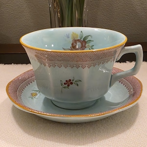 May include: A light blue teacup and saucer set. The cup and saucer have a yellow trim and a decorative brown band. Floral designs adorn the cup and saucer. The teacup has a square handle.