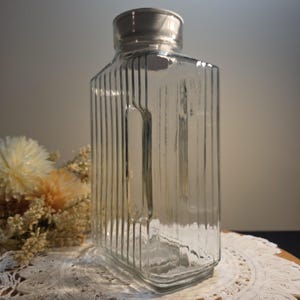 May include: A clear, rectangular glass bottle with vertical ribbed detailing and a silver-colored lid. The bottle is displayed on a white lace doily, with dried flowers in the background. The bottle's design is simple and elegant.