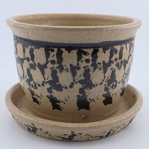 May include: A beige ceramic plant pot with a speckled texture and a dark brown, abstract sponge-painted pattern around the exterior. The pot features a wide, flared rim and sits on a matching circular ceramic saucer with a similar speckled finish.