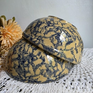 May include: Two stacked stoneware bowls with a tan base and a blue, spatter-like pattern. The bowls are round and have a slightly flared rim. The bowls are sitting on a white lace tablecloth.
