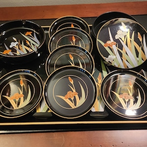 May include: A black lacquerware serving set featuring a tray, plates, and a covered dish. Each piece is decorated with a floral design in orange, white, and gold. The set includes an ashtray. A classic Japanese design.