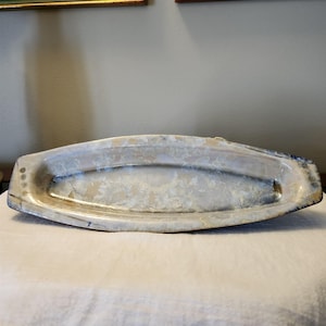 May include: A long, oval-shaped ceramic serving tray with a light beige and blue-grey speckled pattern. The tray has a slightly raised rim and is ideal for serving appetizers or displaying decorative items.