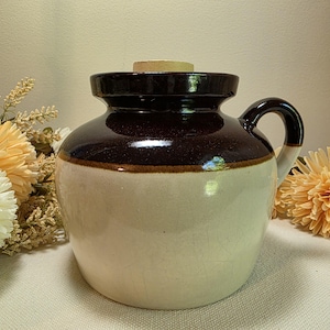May include: A ceramic crock featuring a two-tone design with a dark brown glazed upper section and a cream-colored lower body. The vessel includes a single side handle, a wide mouth, and a flat, light-colored wooden lid resting on the top rim.