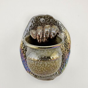 May include: A wall-mounted ceramic vessel featuring a sculpted hand gripping the rim. The piece has a mottled, metallic glaze with iridescent blue, gold, and brown tones. The back plate is oval-shaped with a textured surface and a hole for hanging.
