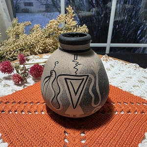 May include: A handcrafted, round pottery vase with a black rim and base. The vase features a beige background with black geometric and abstract designs. The vase is displayed on an orange crocheted doily.